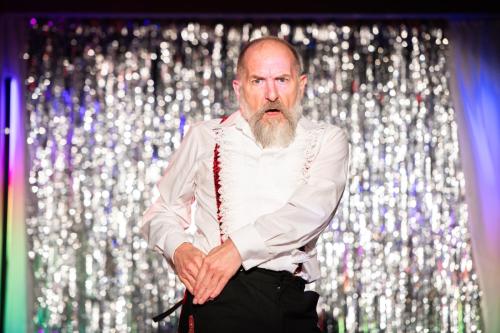 Stephen Docherty on stage in front of a silver shimmering curtain.  